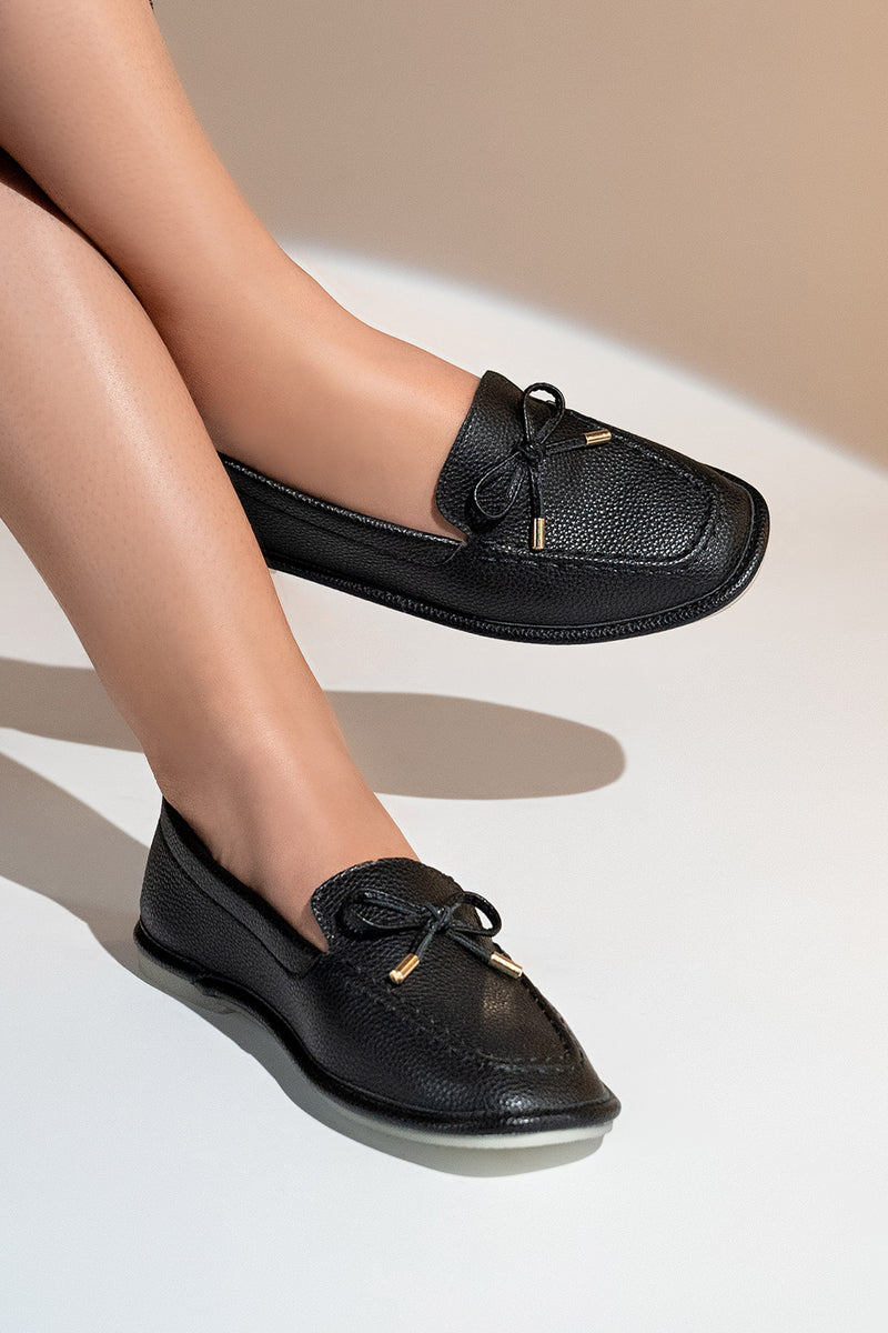 Women's flat moccasin style shoes in black colour and leather texture with matching bow and flexible transparent rubber sole by JULKE. 