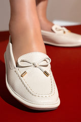 Women's flat moccasin style shoes in off white colour and leather texture with matching bow and flexible transparent rubber sole by JULKE. 